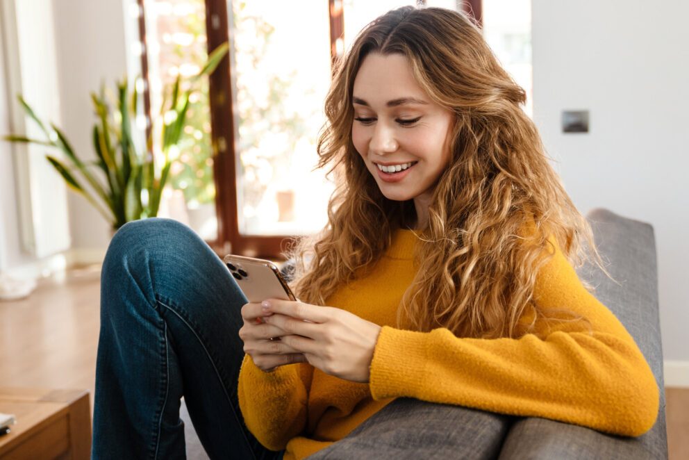 Happy beautiful girl smiling and using cellphone while resting on couch